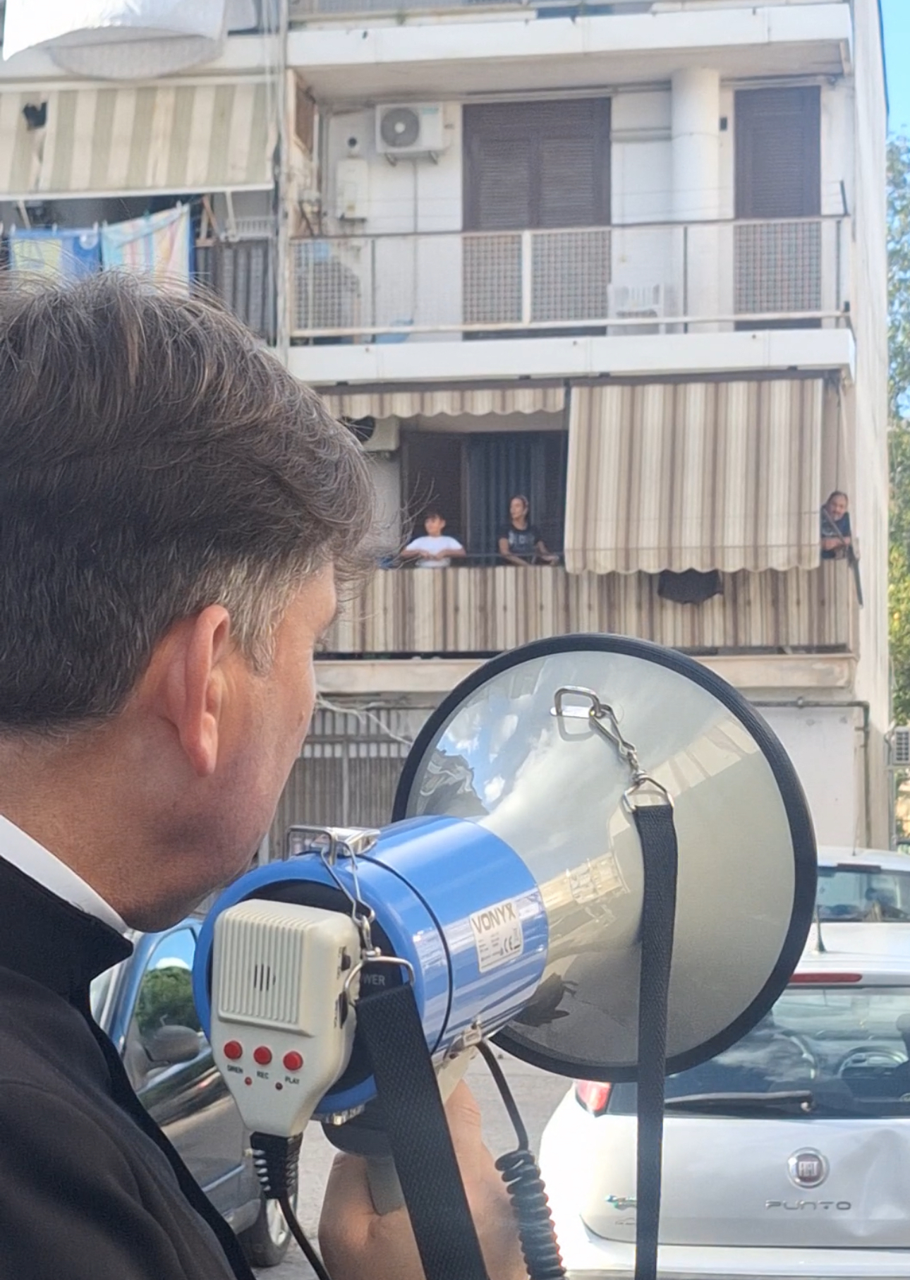 Don Antonio Coluccia parla con il megafono ai residenti affacciati ai balconi di Pianura
